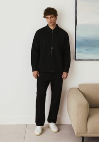 Young man with curly hair wearing a black zip-up jacket, black pants, and white sneakers, standing near a beige sofa and a blue painting.