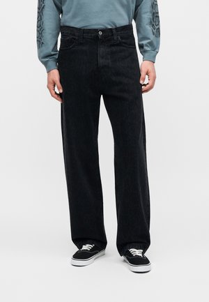 Muž nosiaci voľné čierne rifle, čierno-biele tenisky a modrú mikinu so vzorovanými rukávmi, stojaci na jednofarebnom pozadí.