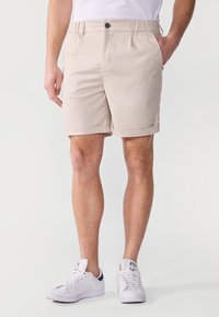 Shorts beiges avec une texture lisse, dotés d'une fermeture à bouton, de poches latérales et de revers roulés. Portés avec des baskets blanches.