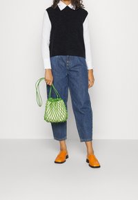 Svart ärmlös tröja över vit skjorta med krage, blå smala jeans, orangea slip-on-skor och en grön nätväska.