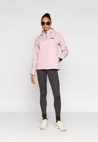 Ανοιχτό ροζ αδιάβροχο μπουφάν με κουκούλα, σχεδιασμένο με εμπρός φερμουάρ και πλαϊνές τσέπες. Συνδυασμένο με σκούρο κολάν και ανοιχτά αθλητικά παπούτσια.