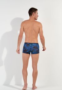Herren-Badehosen mit einem blauen tropischen Blättermuster, schwarzem Bund und einer figurbetonten Passform, von hinten vor einem schlichten Hintergrund gezeigt.
