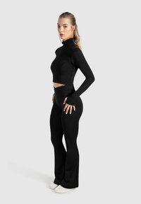 Ensemble de sport noir comprenant un haut cropped à manches longues avec un col montant et des leggings évasés. Texture lisse, design ajusté et discret détail de fermeture éclair.