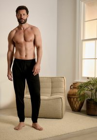 Pantaloni negri de relaxare cu un brâu elastic, purtați de un bărbat cu trunchiul gol. Fundalul include o canapea bej și plante decorative.