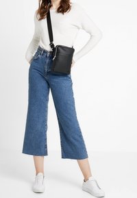 Svart läder crossbody-väska med dragkedjestängning, platt framficka och justerbar rem. Modellen bär en vit långärmad topp, vida jeans och vita sneakers.