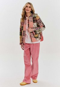 Barevná kostkovaná bunda s květinovými vzory, v kombinaci s růžovými manšestrovými kalhotami a žlutými botami. Model stojí na neutrálním pozadí.