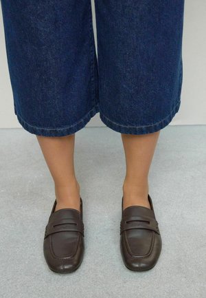 Mocassins en cuir marron avec un bout carré et une bande décorative. Portés avec des culottes en denim larges et sombres, mettant en valeur un dessus lisse et des détails cousus.