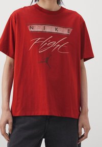 Osoba má na sobě červené tričko Nike s grafikou "Flight" a siluetou skákajícího sportovce, které doplňují tmavé kalhoty.