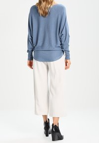 Hellblaues, übergroßes Sweatshirt mit abgerundetem Saum, kombiniert mit weißen, weit geschnittenen Hosen. Schwarze, hochhackige Stiefeletten runden den Look ab.