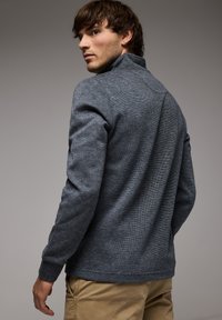 Pullover grigio testurizzato con colletto alto, maniche lunghe e polsini a coste. Il tessuto presenta un motivo sottile, che completa i pantaloni color sabbia.