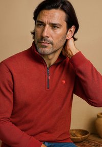 Rode pullover met ritssluiting, gemaakt van een gestructureerde stof, met een halve rits en een klein logo op de borst. De achtergrond is neutraal.