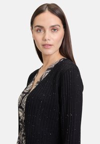 Cardigan noir côtelé avec des détails scintillants, décolleté en V et manches longues. Porté par-dessus un haut à motifs. Texture lisse.