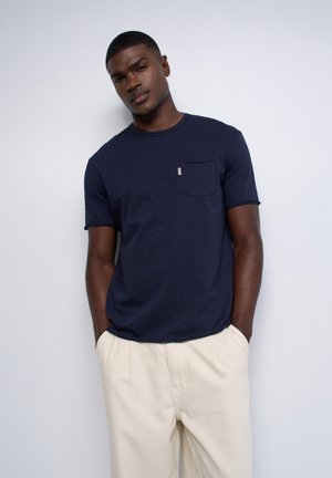 Homme portant un t-shirt bleu marine à manches courtes avec poche poitrine et pantalon beige clair, debout contre un mur blanc uni.