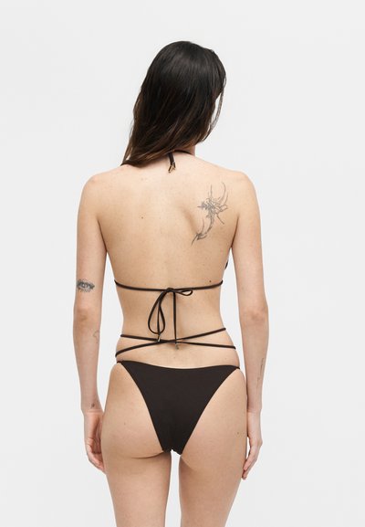 Γυναίκα με μακριά σκούρα μαλλιά που φοράει μαύρο στρινγκ μπικίνι, δείχνοντας την πλάτη με τατουάζ λουλουδιών και ματιών στο αριστερό χέρι και στη δεξιά ωμοπλάτη.