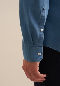 Primo piano di una mano accanto a una manica di camicia blu con un piccolo ricamo di rosa vicino al polsino.