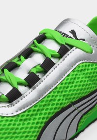 Baskets Puma argentées et vertes avec côtés en mesh, lacets vert néon passés dans des boucles noires, et bande logo argentée cousue.