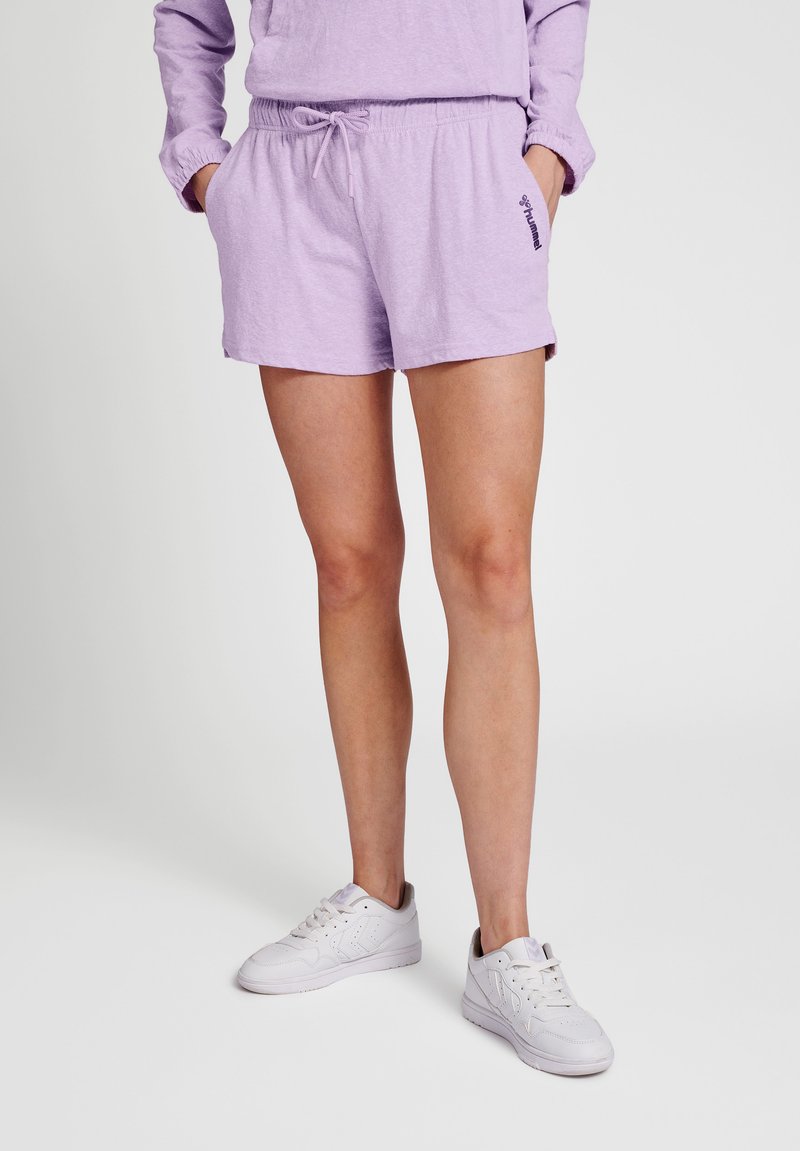 Personne portant un short à cordon violet pastel et un sweat assorti, mains dans les poches, debout en baskets blanches sur fond uni.