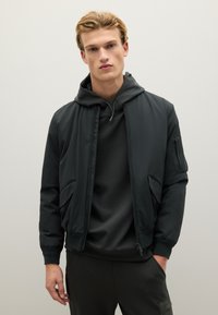 Zwarte bomberjack van glad, lichtgewicht materiaal, met een ritssluiting aan de voorkant, zijzakken en een hoodie eronder.