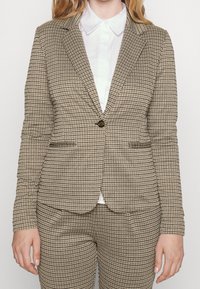Blazer à carreaux marron et crème avec une fermeture à un bouton, des poches latérales et une coupe ajustée, associé à une chemise blanche à col en dessous.