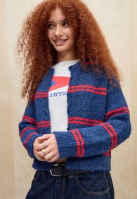 Jonge vrouw met krullend rood haar, gekleed in een blauwe cardigan met rode strepen, een wit T-shirt met een opdruk, donkere jeans en meerdere ringen om haar vingers.