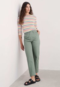 Un haut à manches longues rayé présente des lignes rouges, vertes et jaunes. Un pantalon taille haute vert et des sandales noires avec un détail de boucle. Textures lisses.