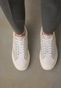 Sneakers bianchi e beige con tomaia testurizzata, lacci piatti e suola spessa. Gli accenti laterali presentano una sottile linea blu.