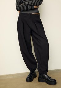 Čierne plisované nohavice s priliehavým strihom a elastickými manžetami, skombinované s robustnými čiernymi šnurovacími topánkami. Dlhý rukáv s horizontálnym pruhovaním.