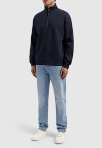 Navy blue zip-neck sweatshirt in soft fabric, featuring ribbed cuffs and hem, paired with light blue jeans and white sneakers.