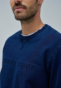 Sudadera azul marino hecha de tejido texturizado que presenta un logo en relieve. Tiene un cuello redondo con puños y dobladillo de canalé flexibles.