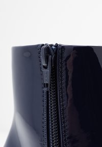 Botín de charol azul marino con cremallera lateral y marca YKK. Presenta una textura suave y brillante, además de una forma ajustada y contorneada.