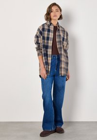 Plaid shirt in beige, navy, and brown; button-up design. Brown top, high-waisted blue flared jeans, and dark brown footwear.