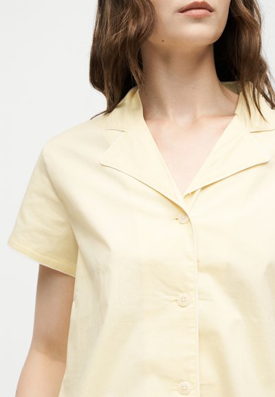 Femme portant une chemise jaune pâle à manches courtes boutonnée avec col à revers et cheveux clairs ondulés, coupés au niveau du menton.