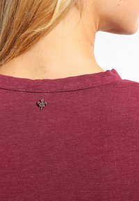 Bordeauxfarbenes Baumwoll-T-Shirt mit Rundhalsausschnitt; verfügt über eine kleine schwarze Ahornblattnadel auf der Rückseite, hat kurze Ärmel und eine glatte Textur.