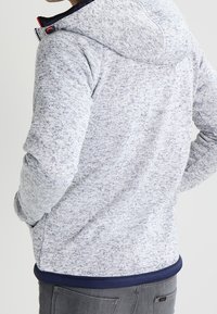 Person som bär en ljusgråmelerad huva jacka med händerna i fickorna, sedd bakifrån.