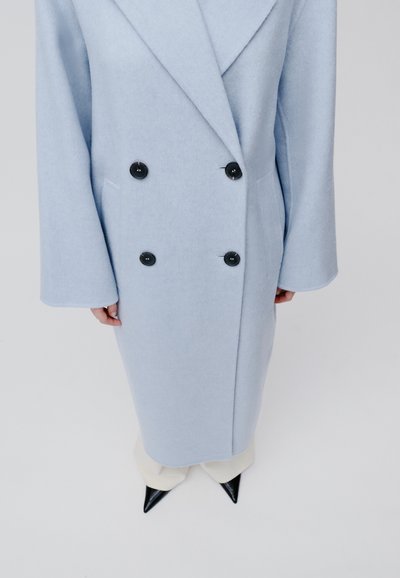 Cappotto blu doppiopetto realizzato in tessuto morbido, con bottoni neri, rever oversize e maniche ampie con silhouette dritta.