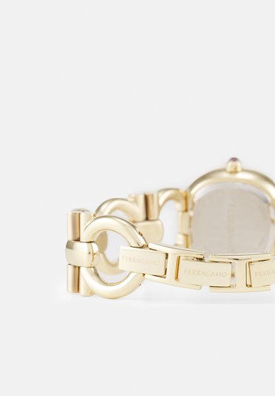 Gold metal watch with a round case and linked bracelet design. Features engraved accents on links and a small gem at the crown.
