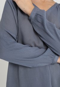 Blouse grise texturée à manches longues avec poignets boutonnés et décolleté en V, mettant en avant un tissu doux et léger.