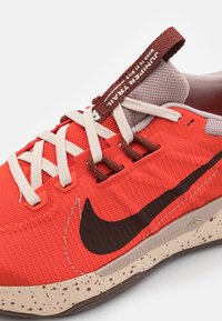Scarpa sportiva rossa con tomaia in rete, lacci beige e logo swoosh nero. Suola intermedia beige con macchie evidenti e una linguetta intrecciata sul tallone.