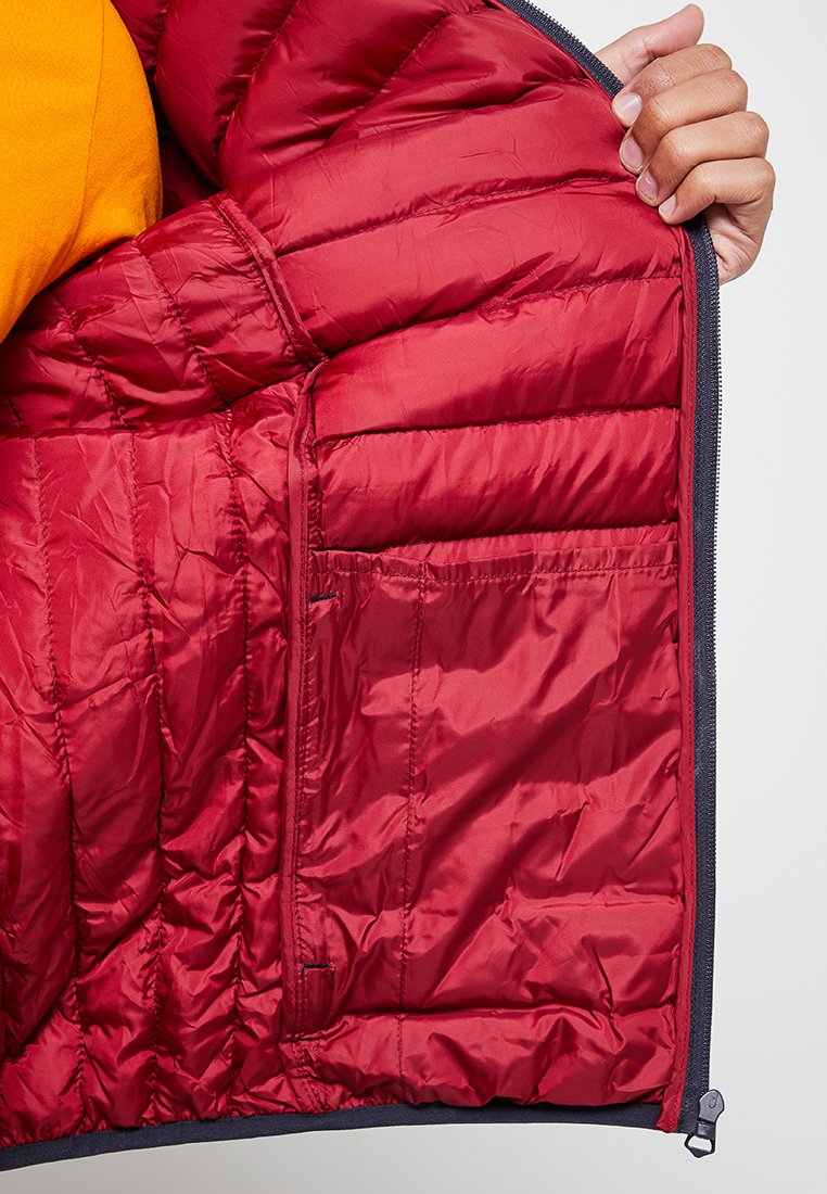 Eine Hand hält eine offene rote gesteppte Jacke, die die Innenstiche und die Innentasche vor einem grauen Hintergrund zeigt.