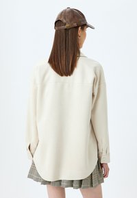 Beige långärmad skjorta med en rundad nederkant och knappmanchetter, i kombination med en rutmönstrad plisserad kjol och en brun läderkeps.