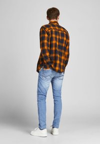 Man som står med ryggen mot kameran, iförd en orange- och svart-rutigt flanellskjorta, ljusblå jeans och vita sneakers, mot en enhetlig grå bakgrund.