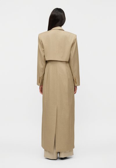 Γυναίκα με μακριά σκούρα μαλλιά που φοράει μακρύ μπεζ παλτό και παντελόνια με φαρδιά μπατζάκια, όρθια με την πλάτη στραμμένη προς ένα απλό φόντο.