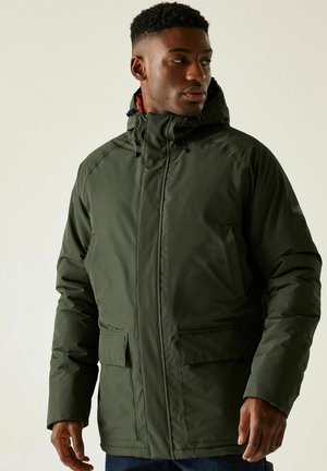 Mężczyzna w zielonej zimowej kurtce z kapturem i przednimi kieszeniami, patrzący w prawo na jednolitym jasnym tle.