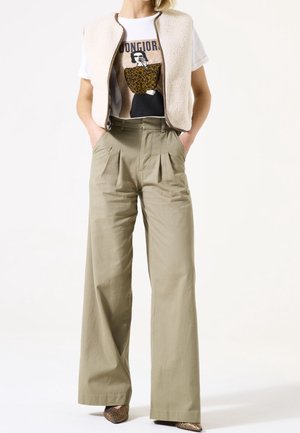 Femme portant une veste beige en shearling sur un t-shirt graphique, un pantalon kaki taille haute à jambes larges et des chaussures pointues en peau de serpent, mains dans les poches.