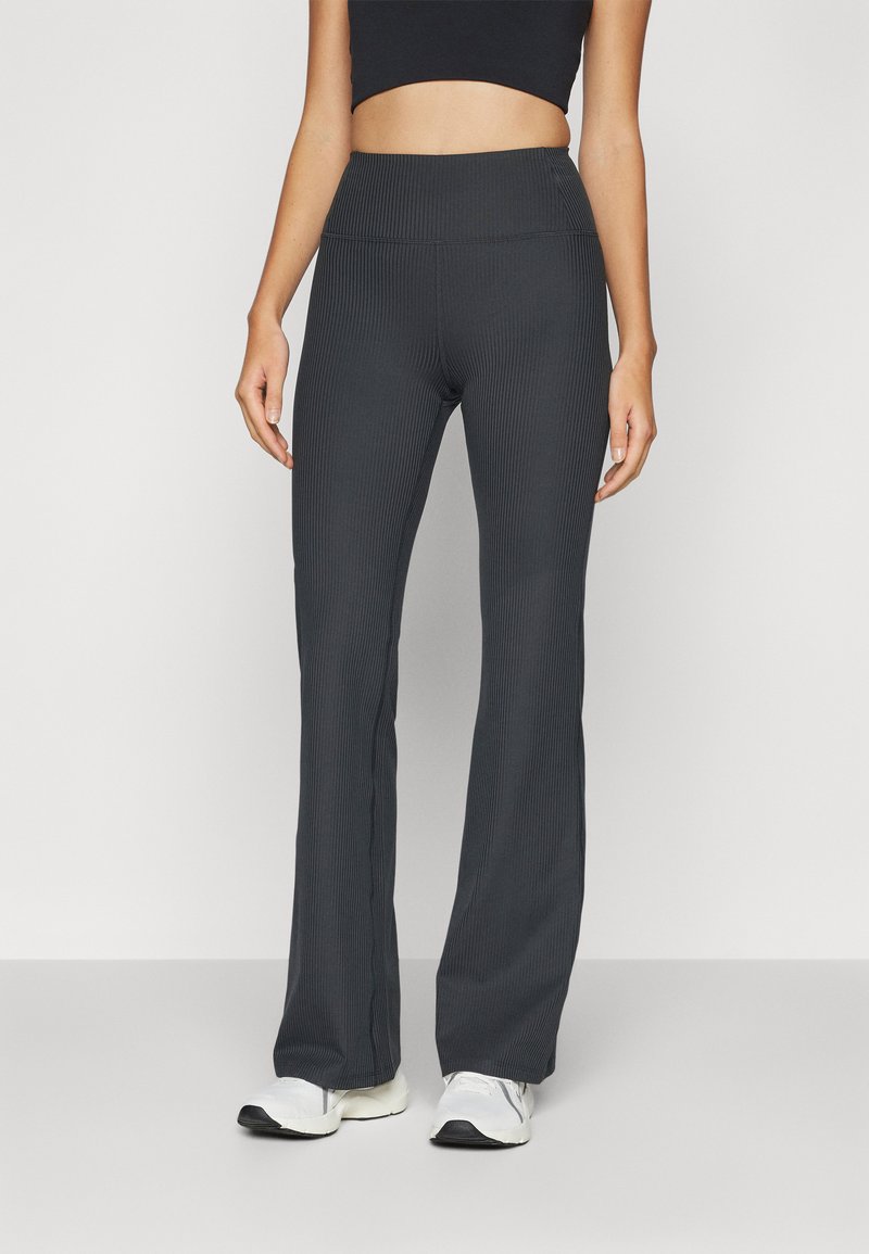 Pantalons de yoga gris foncé à côtes avec une taille haute et un ourlet évasé, associés à des chaussures de sport blanches. Texture lisse, design décontracté.