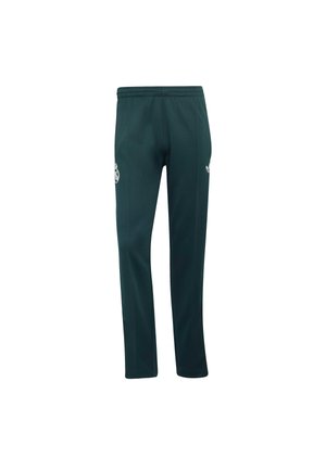 Personne portant un pantalon de sport vert foncé avec des bandes blanches sur les côtés et un logo, des baskets blanches et une chemise de couleur claire, debout les mains dans les poches.