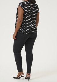 Blouse noire à pois avec des manches courtes, en tissu doux, détail de bouton dans le dos ; assortie avec un jean noir ajusté et des chaussures à talons hauts noires.