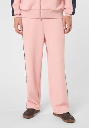 Parte inferior del cuerpo de una persona que lleva pantalones sueltos rosas con rayas negras laterales, chaqueta rosa y zapatos marrón oscuro sobre un fondo liso.