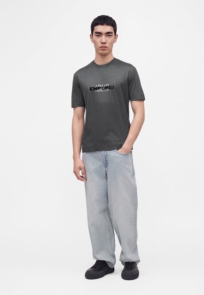 Jeune homme debout portant un t-shirt graphique gris foncé, un jean ample bleu clair et des baskets noires sur un fond blanc uni.