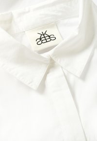 Bela bombažna srajca s koničastim ovratnikom, sprednjim gumbnim delom in tkaninsko oznako z črnim logotipom, ušito znotraj ovratnika.